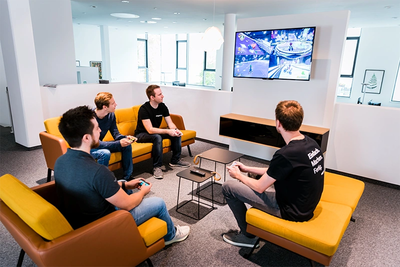 Vier Personen sitzen auf gelben Sofas in einem modernen Büro und spielen gemeinsam Videospiele auf einem großen Bildschirm an der Wand.