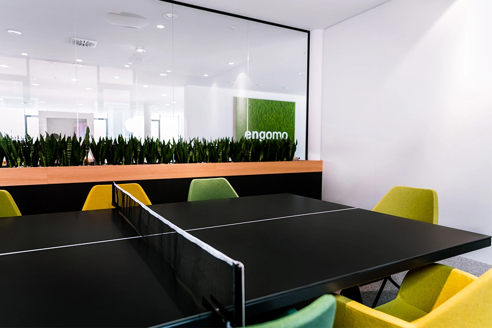 A modern office space featuring a ping pong table surrounded by yellow-green chairs and a divider with plants; the “engomo” logo is visible in the background.