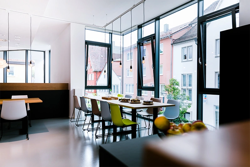 Ein moderner Essbereich im engomo-Office mit großen Fenstern, einem langen Tisch, weißen und grünen Stühlen sowie Tellern mit Kuchen.