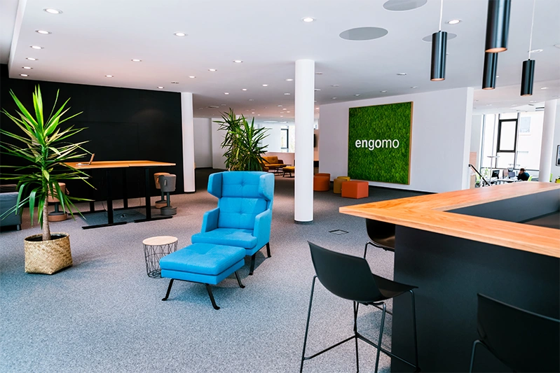 The image shows the modern engomo office with a striking blue chair in the center, green plants, and a cozy open-space design.