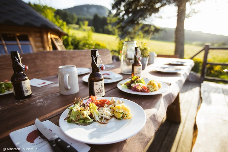 A wooden outdoor table set with plates of regional food, beer bottles, and mugs, surrounded by scenic countryside at sunset.
