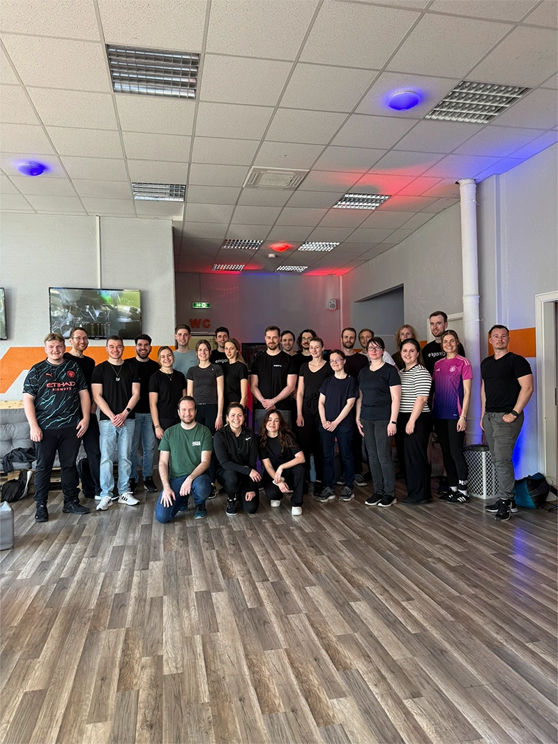 Eine größere Gruppe von Menschen steht und kniet gemeinsam in einem Raum mit Holzboden und Bildschirmen an den Wänden, für ein Gruppenfoto.