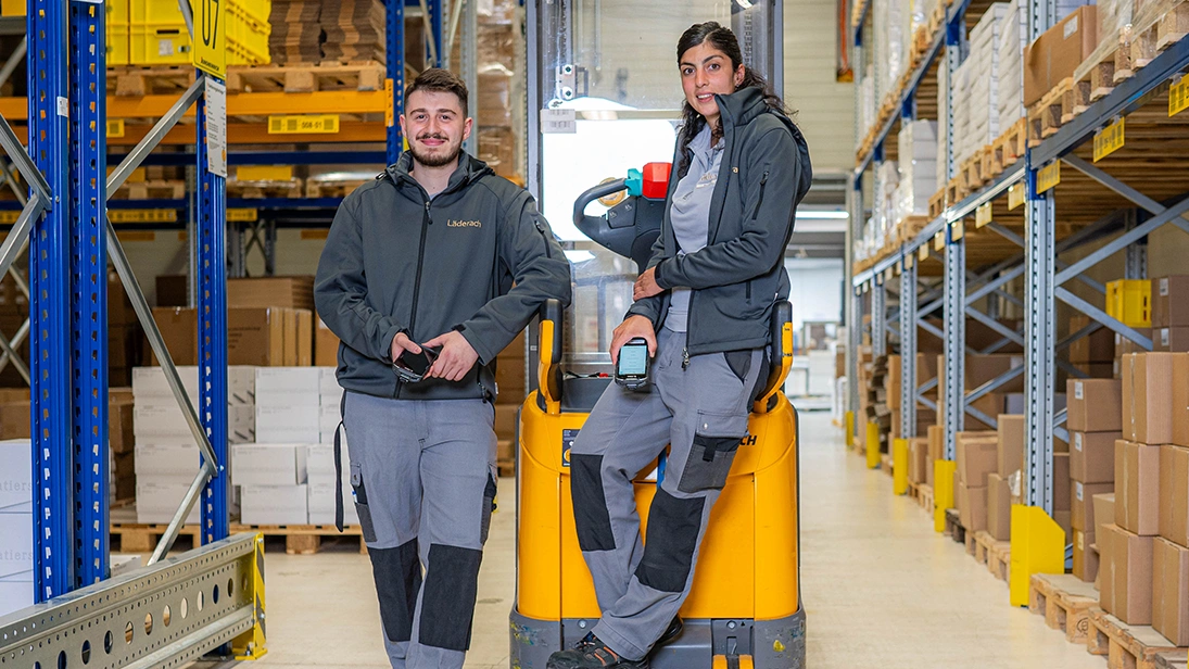 The image shows the interior of a warehouse from Läderach. In the center, there is a forklift with one person leaning on it and another sitting in it.