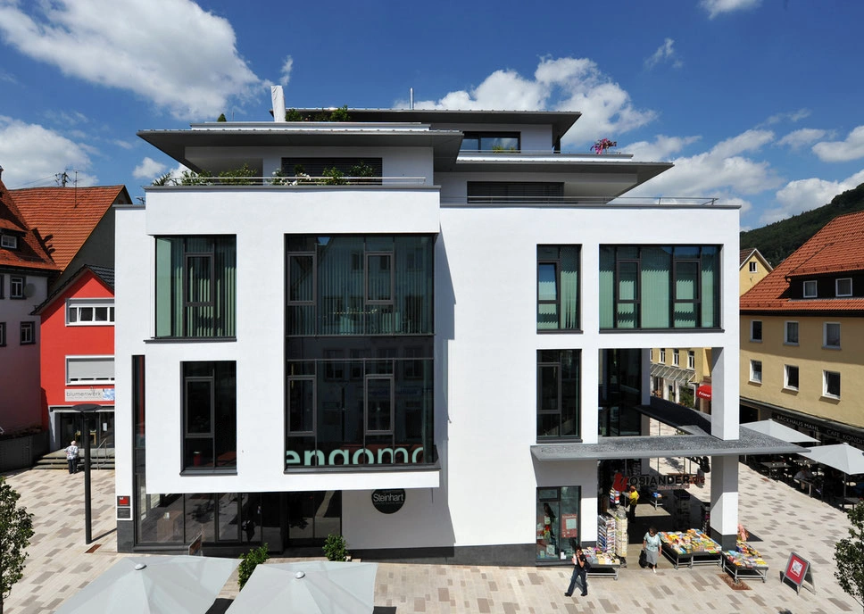 Modern office building housing engomo’s headquarters in downtown Albstadt, featuring large glass facades and surrounding retail stores on a sunny day.
