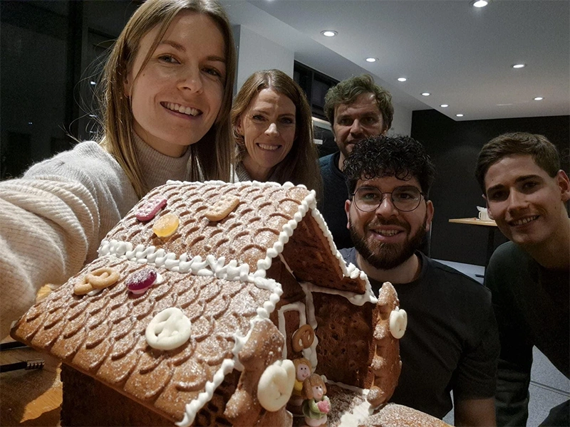 Five people smiling at the camera while presenting a detailed gingerbread house in a cozy indoor setting.