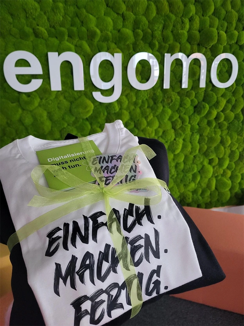Ein gefaltetes T-Shirt mit der Aufschrift „Einfach. Machen. Fertig.“ ist mit einer grünen Schleife verpackt und liegt vor einer grünen Mooswand mit dem engomo-Logo.