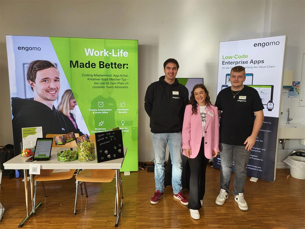 Drei Personen stehen vor einem Messestand mit grünen und weißen Bannern des Unternehmens „engomo“. Auf dem Tisch liegen Flyer, ein Tablet und Schalen mit Süßigkeiten.
