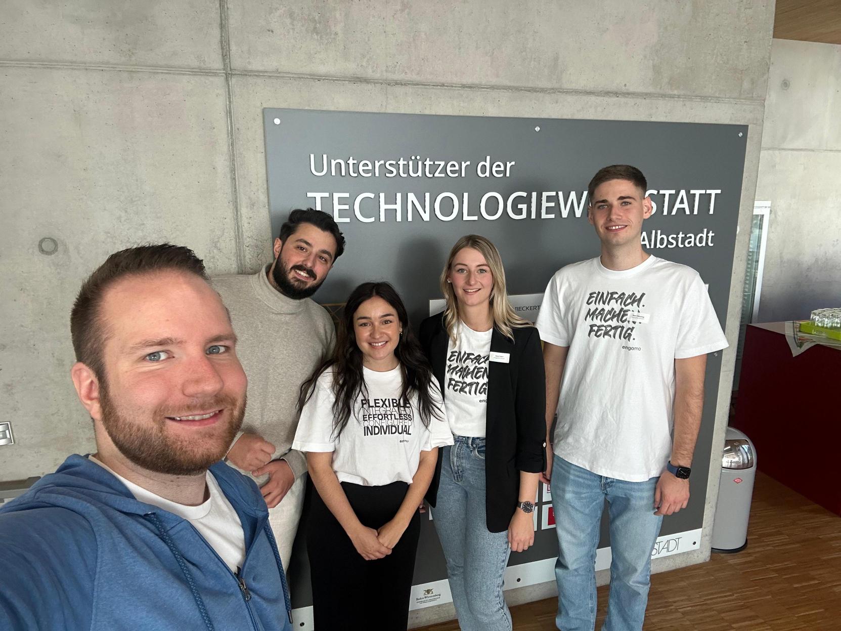 Fünf Personen stehen lächelnd vor einer Wand mit dem Schriftzug „Unterstützer der Technologiewerkstatt Albstadt“ und tragen engomo-Shirts.