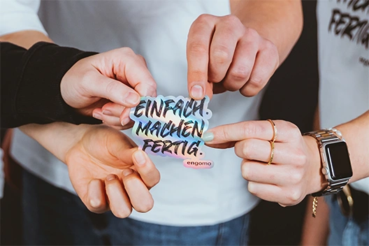 The image shows four hands holding a sticker with the words