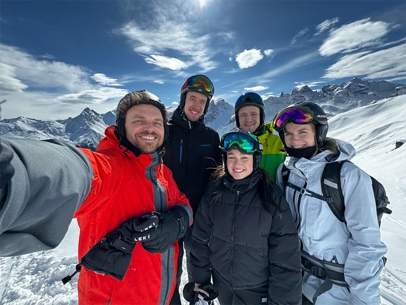 A group of five people in ski gear smiling in the snow with a stunning mountain backdrop under a clear blue sky.