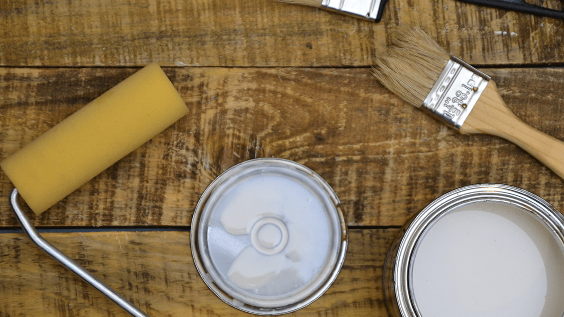 Open cans of white paint, a paintbrush, and a paint roller placed on a rustic wooden surface – ready for painting.
