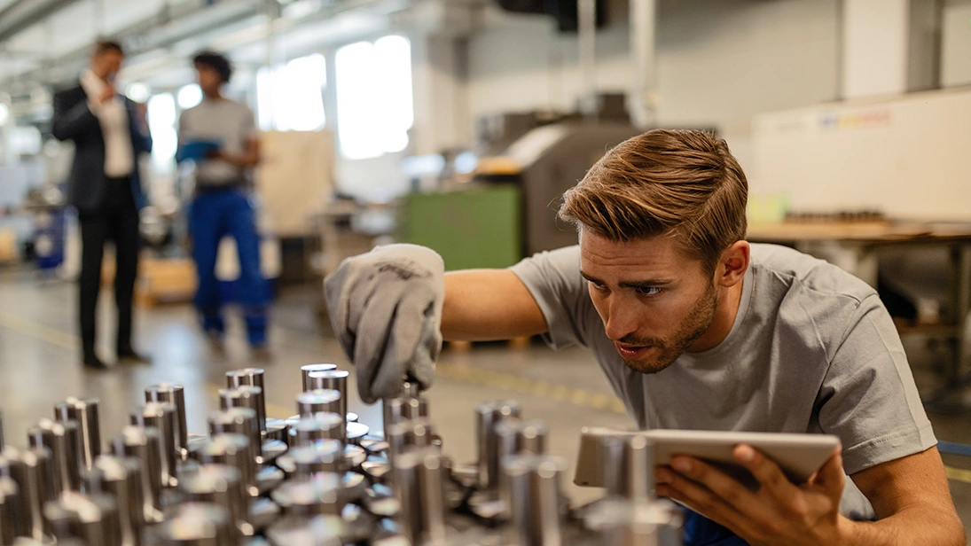 Mitarbeiter prüft in einer Produktionshalle Bauteile auf einem Tisch und hält dabei ein Tablet – symbolisiert digitale Qualitätssicherung in der Fertigung.