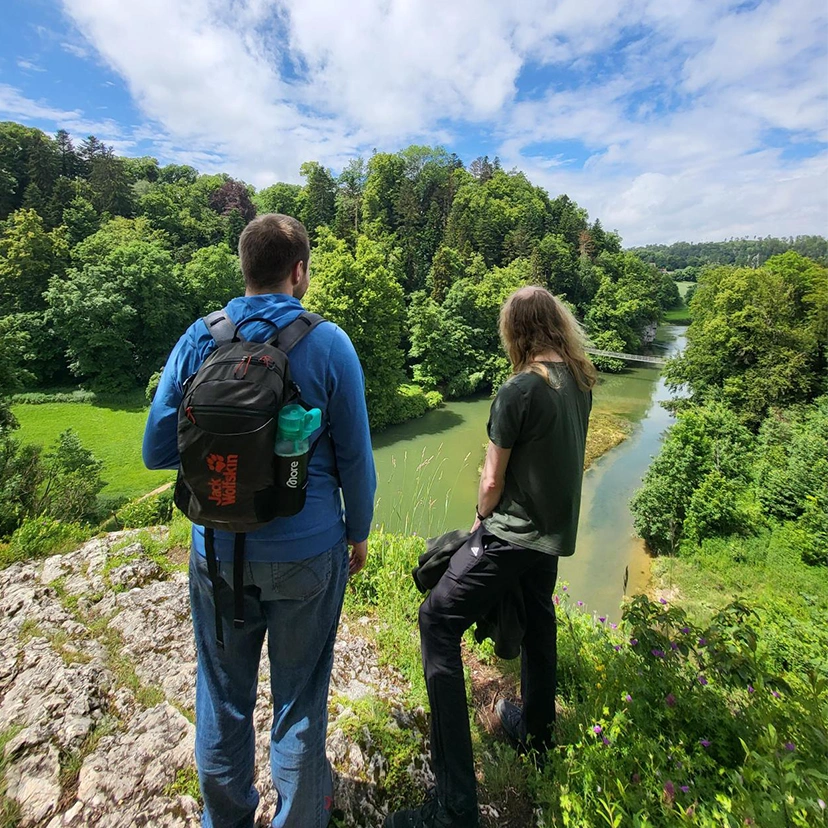 Two people with hiking gear are standing at the edge of a valley, seen from behind, looking down