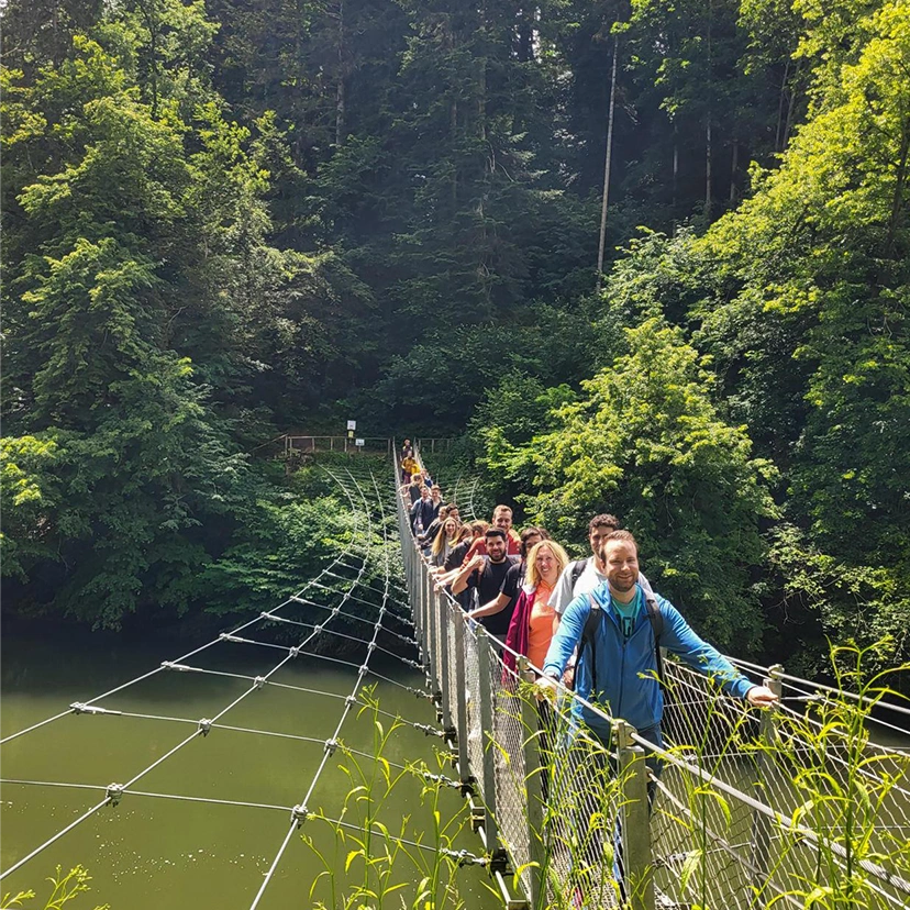 A group of people smiling while standing on a suspension bridge over a green river, surrounded by dense forest and summer sunlight.