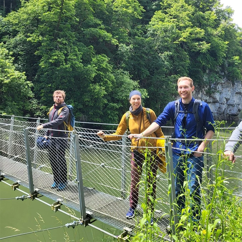 Three hikers smiling while standing on a suspension bridge over a river, surrounded by dense green forest.
