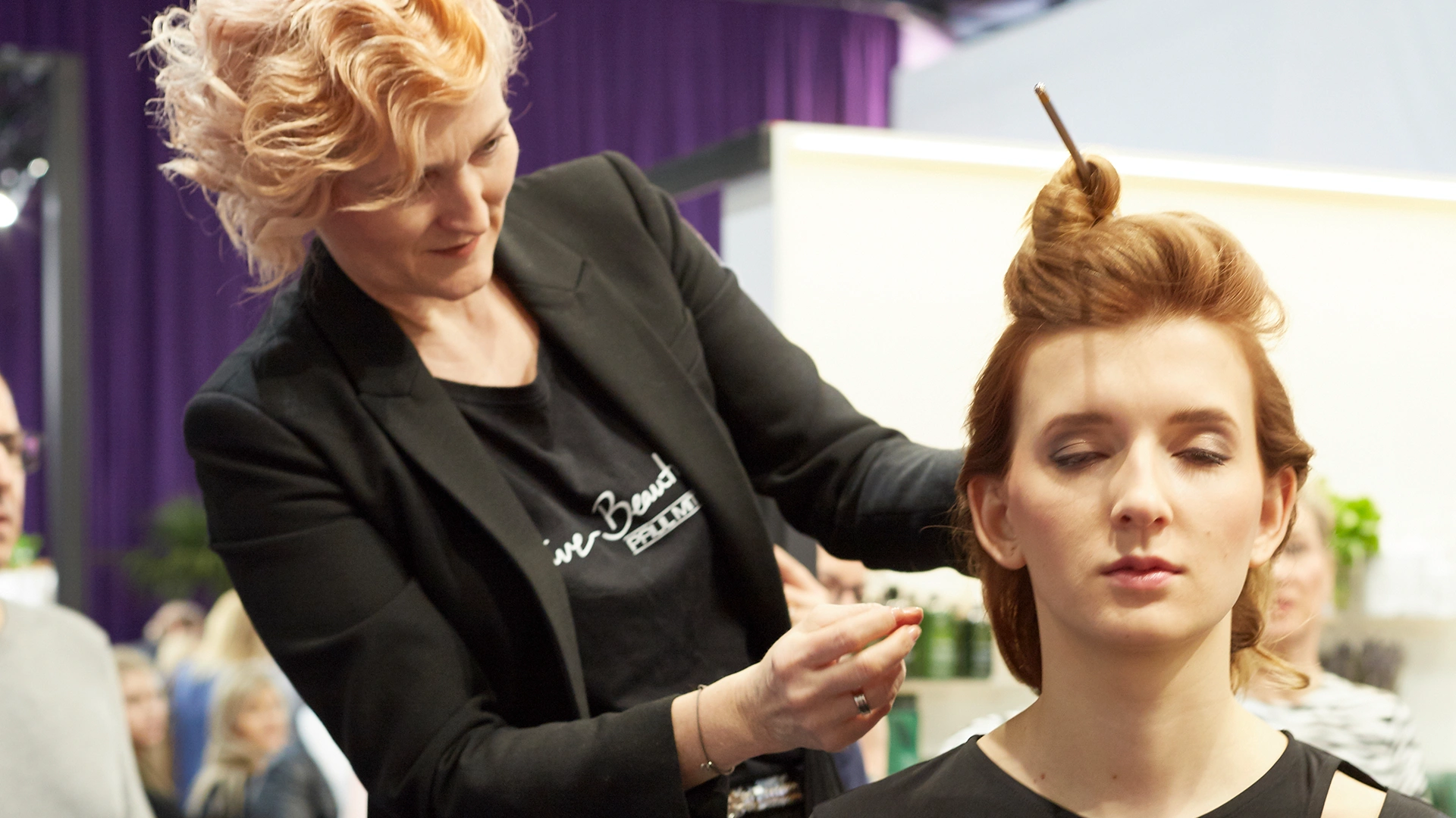 Hairdresser styling a young woman’s hair, who has her eyes closed, during a trade fair or event. Other visitors and an exhibition booth can be seen in the background.