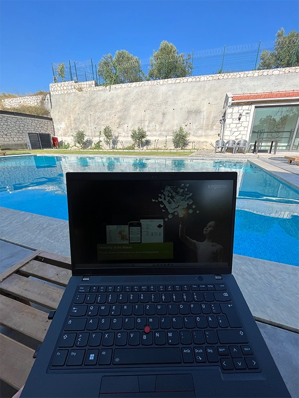 Ein geöffneter Laptop mit einer engomo-Präsentation steht auf einem Tisch am Pool unter blauem Himmel – Arbeit im Freien.