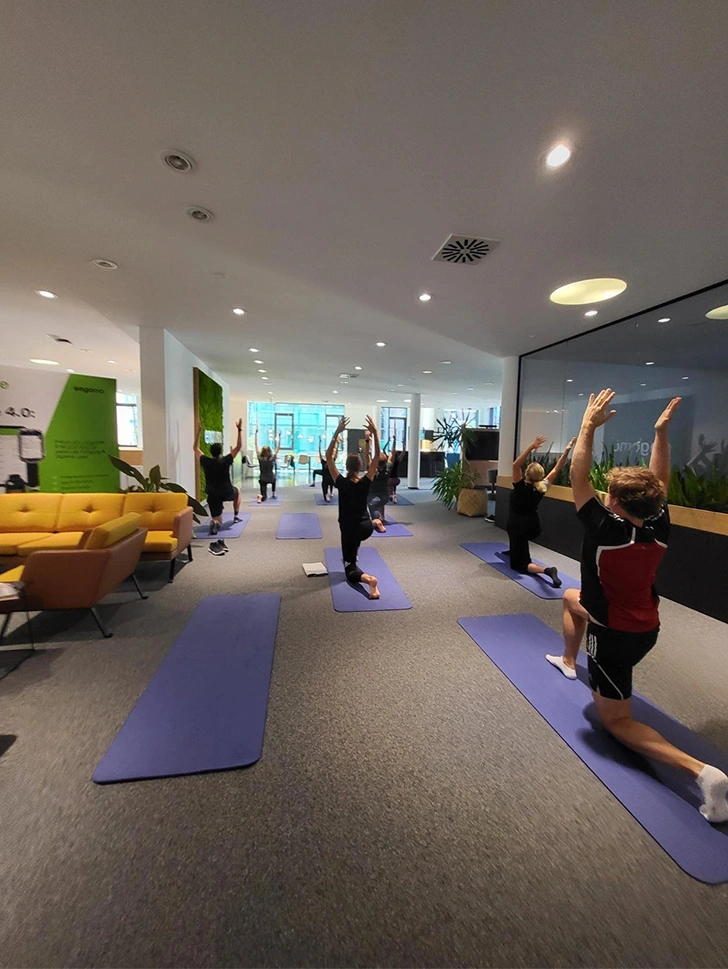 Mehrere Personen machen Yogaübungen auf blauen Matten in einem modernen Büro mit großen Fenstern, Pflanzen und Sitzgelegenheiten.