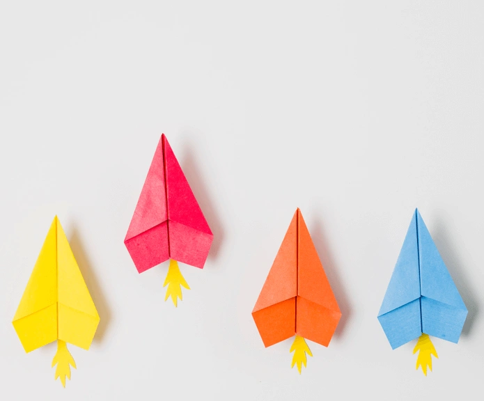 Vier bunte Papier-Raketen in Gelb, Rot, Orange und Blau auf weißem Hintergrund – symbolisieren kreative Vielfalt, Aufstieg und individuelle Lösungen.