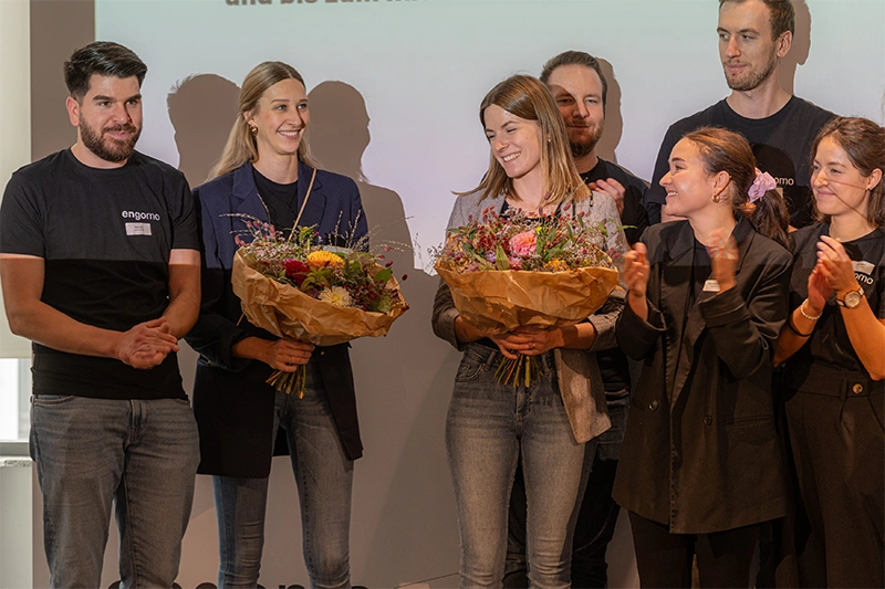 Several people standing on a stage; two women in the center holding flower bouquets being applauded by their colleagues.