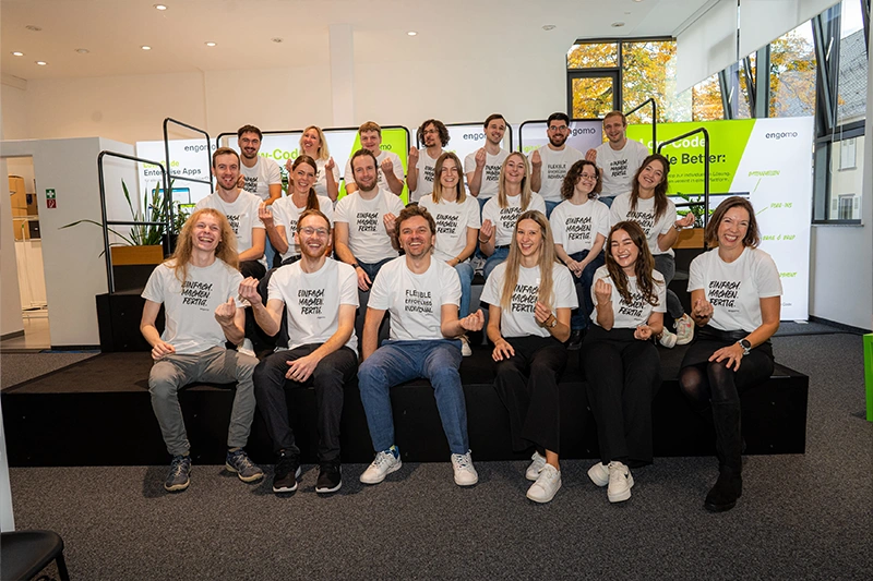 Eine fröhliche Gruppe von Menschen hebt lachend die Arme auf einem Messestand von engomo, umgeben von bunten Messeständen und Bildschirmen.