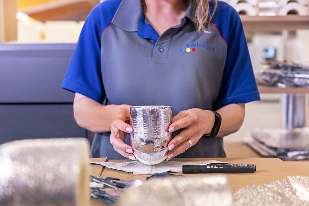 Eine Mitarbeiterin von swisspor h&auml;lt ein mit Aluminiumfolie umwickeltes D&auml;mmstoffst&uuml;ck in den H&auml;nden und bereitet es an einem Arbeitstisch in der Produktion weiter vor.