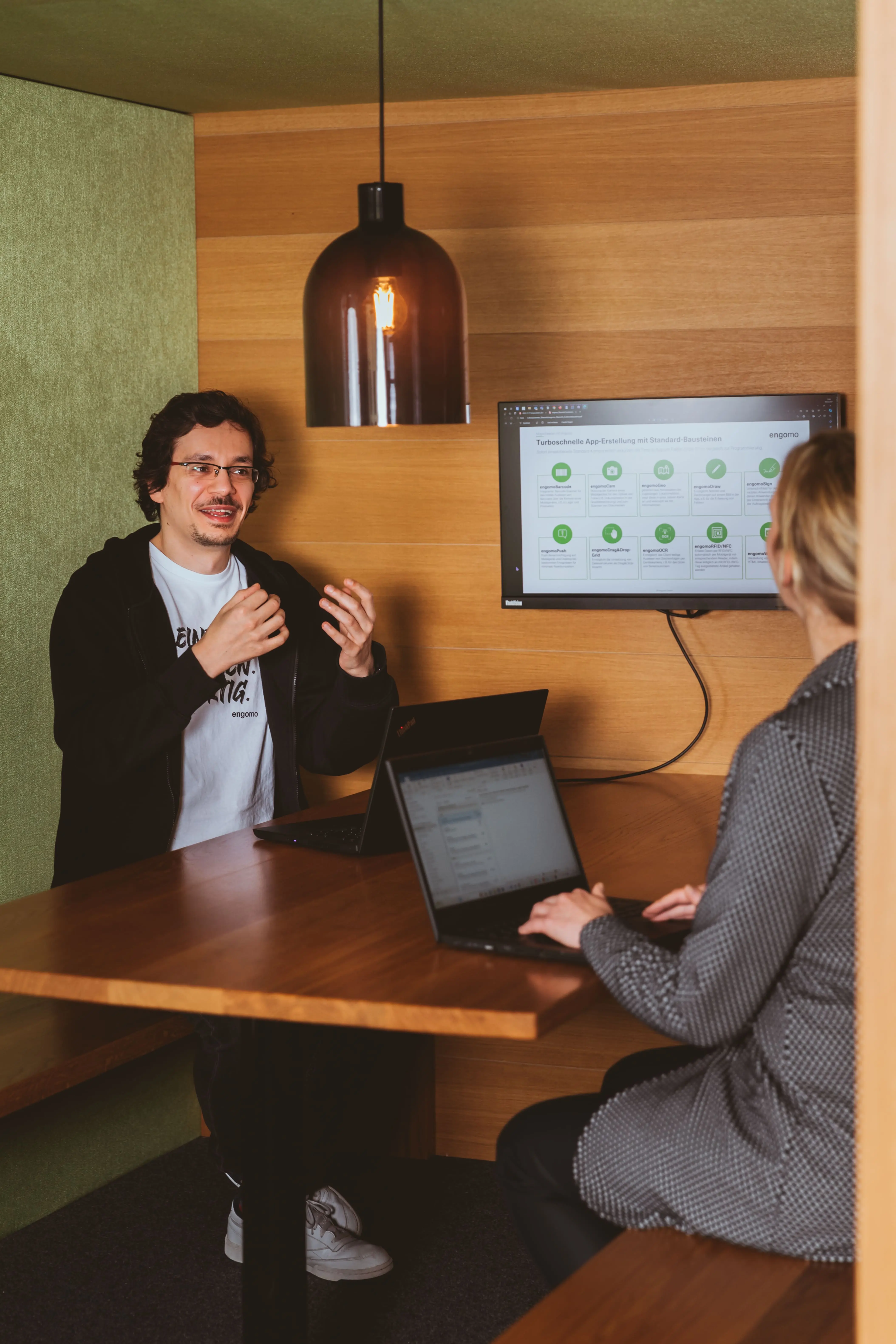 Eine von hinten dargestellte Person schaut auf eine andere Person, die vor einem Bildschirm steht und mit Gesten spricht.