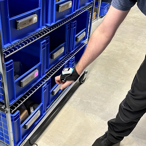 Employee at KIND GmbH & Co. KG scans storage bins using a wearable scanning device between blue containers on a rolling shelf.