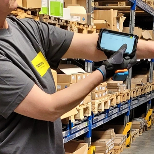 Employee wearing an Ammon shirt and gloves uses a mobile scanner to capture data in front of warehouse shelves stocked with fittings.
