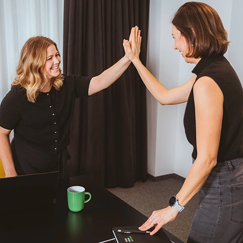 Two people giving each other a high-five in a modern office – symbolizing teamwork, success, and a “just do it!” mindset.