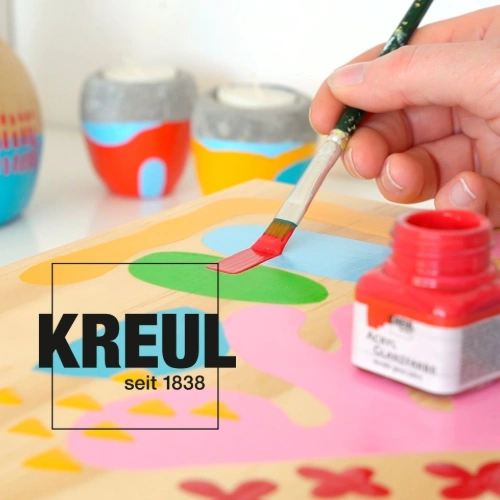 A hand painting a colorful wooden surface with a brush next to an open red paint jar by KREUL, with decorated vessels in the background.