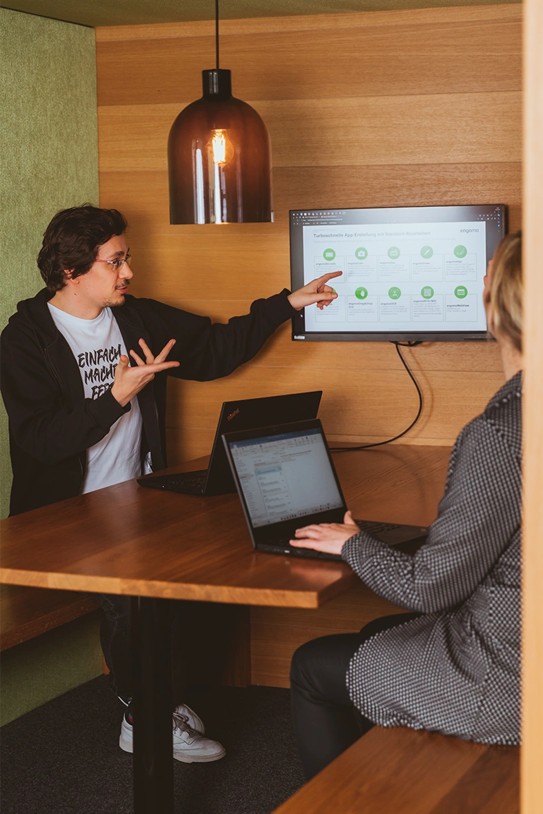 Two people sitting in front of a screen, with the person on the left pointing at it.