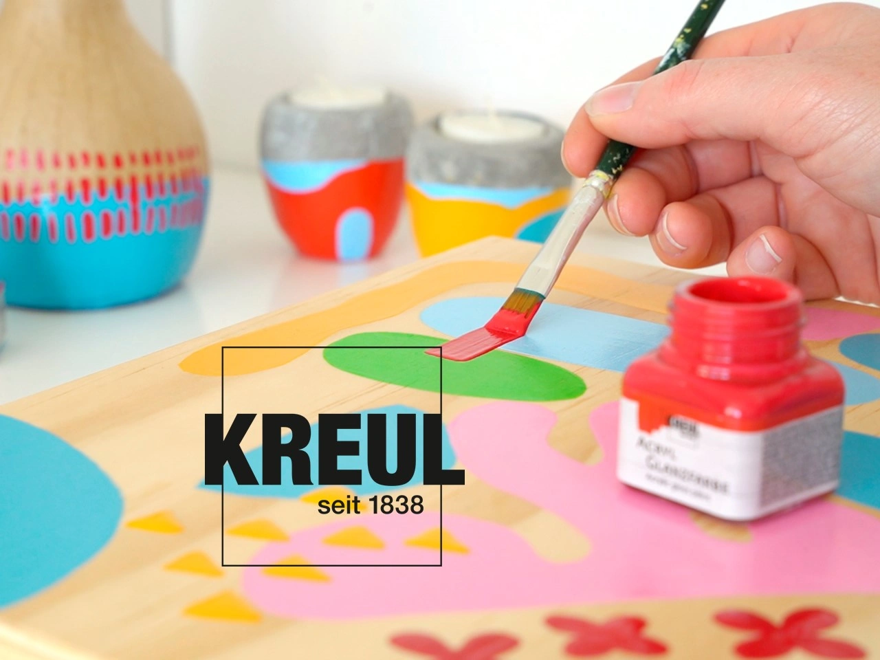 Eine Hand bemalt mit einem Pinsel eine bunte Holzplatte neben einem offenen roten Farbt&ouml;pfchen von KREUL, im Hintergrund stehen dekorierte Gef&auml;&szlig;e.