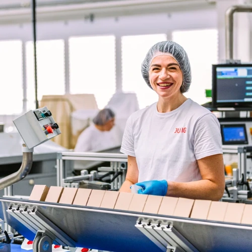 A woman stands at a production line, smiling at the camera. She is wearing a hairnet and gloves. Machines and digital displays can be seen in the background.