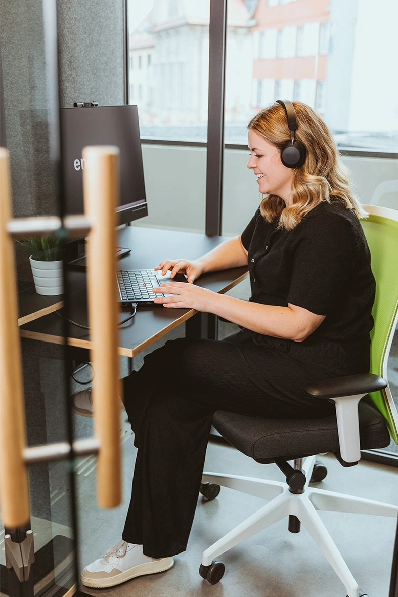 Side view of a woman wearing a headset, sitting in front of a screen and using a laptop’s touchpad.